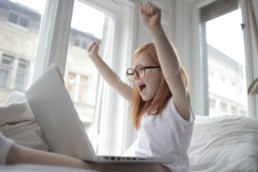 Petite fille qui lève les bras devant son ordinateur en signe de victoire